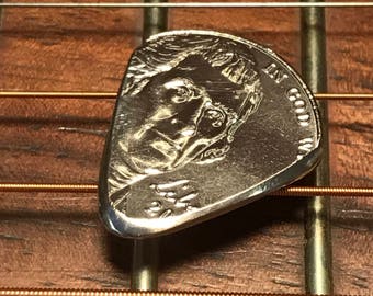 Könnte beinhalten: Ein silberner Gitarrenplektrum in Form einer US-amerikanischen 50-Cent-Münze mit dem Profil eines Mannes auf der Vorderseite und den Worten "In God We Trust" auf der Rückseite.