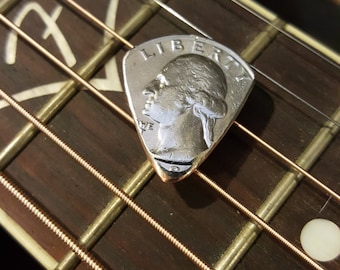Könnte beinhalten: Ein silberner Gitarrenplektrum in Dreiecksform mit den Worten "LIBERTY" und einem Porträt einer Frau. Der Plektrum liegt auf den Saiten einer braunen Akustikgitarre.