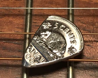 Könnte beinhalten: Ein Gitarrenplektrum aus kanadischem Nickel, mit einem Biber und dem Text "5 CENTS" und "CANADA 2001". Das silberne Plektrum liegt auf Gitarrensaiten und einem hölzernen Griffbrett.
