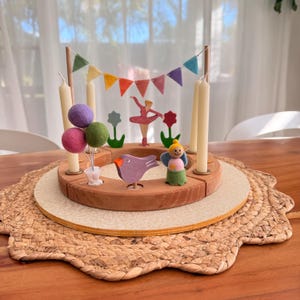 May include: A wooden centrepiece with a circular design, featuring a ballerina, bird, angel, and colourful felt balloons. The centrepiece includes a banner of felt flags and space for candles. The base is set on a woven placemat.