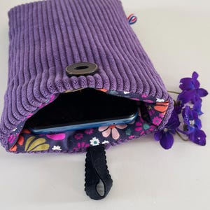 Può includere: Una custodia per telefono in velluto a coste viola con fodera floreale. La custodia ha una chiusura a scatto nera e un passante nero. All'interno è visibile uno smartphone. Un rametto di fiori viola è accanto alla custodia.
