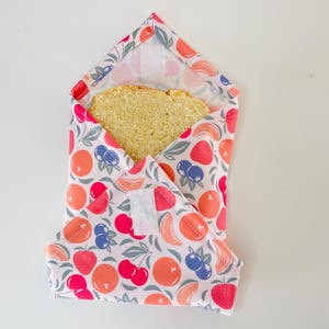 Peut inclure: Un emballage sandwich réutilisable avec un motif de fruits colorés, comprenant des oranges, des fraises, des cerises et des myrtilles. Une tranche de pain est visible à l'intérieur. L'emballage est fermé par un velcro blanc.