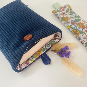 May include: A navy blue corduroy book sleeve with a wooden button fastening. The interior has a floral pattern. A matching floral print bookmark and a small bouquet of dried flowers are also present.