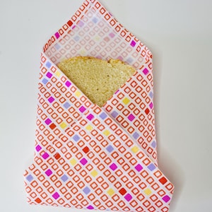 Peut inclure: Une tranche de pain enveloppée dans un emballage sandwich en tissu réutilisable. L'emballage présente un fond blanc avec un motif répétitif de carrés rouges, roses, jaunes et violets. Le pain est brun clair.
