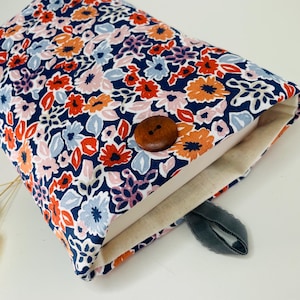 Peut inclure: Une housse de livre à imprimé floral avec un fond bleu marine et un motif de fleurs rouges, oranges, roses et bleues. La housse a une fermeture à bouton en bois et un ruban gris. L'intérieur est beige clair.
