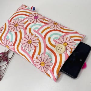 Può includere: Una custodia per telefono floreale con un motivo a margherita in rosa, arancione e bianco. La custodia ha una chiusura con bottone e uno smartphone nero parzialmente visibile all'interno. Un piccolo rametto di fiori secchi si trova nell'angolo in basso a sinistra.