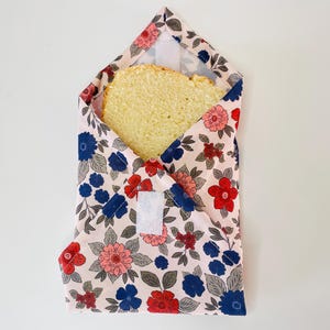 Peut inclure: Un emballage sandwich réutilisable avec un motif floral rose, rouge et bleu. L'emballage est ouvert, révélant une tranche de pain. L'emballage est fixé avec une fermeture velcro blanche. L'emballage est conçu pour contenir des sandwichs.