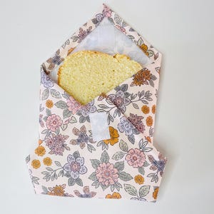 Peut inclure: Un emballage sandwich réutilisable avec un motif floral rose, pêche et lavande. L'emballage est ouvert, révélant une tranche de pain jaune. Une fermeture velcro blanche est visible à l'avant. Conçu pour le stockage des aliments.