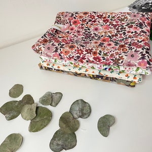 May include: A stack of five fabric squares with floral patterns in shades of pink, red, white, and brown. The fabric squares are folded neatly and are sitting on a white surface. There are several dried green leaves scattered around the fabric squares.