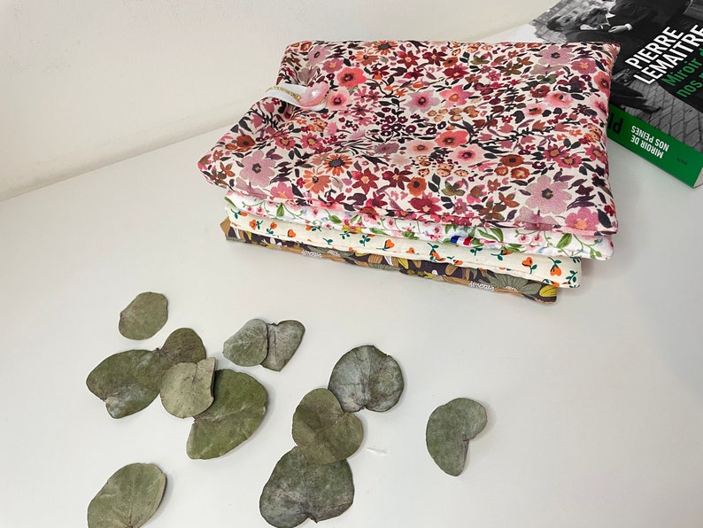 May include: A stack of five fabric squares with floral patterns in shades of pink, red, white, and brown. The fabric squares are folded neatly and are sitting on a white surface. There are several dried green leaves scattered around the fabric squares.