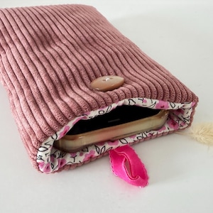 Può includere: Una custodia in velluto a coste color rosa polvere con chiusura a bottone marrone e un passante in tessuto rosa acceso. L'interno è foderato con un tessuto bianco con un motivo di piccoli fiori rosa e foglie nere, contenente uno smartphone.