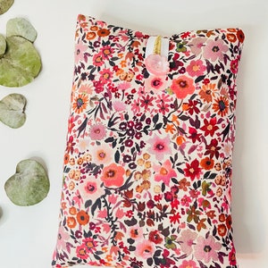 May include: A small, rectangular pillow with a white background and a floral pattern in shades of pink, red, and brown. The pillow has a white ribbon and a pink button.