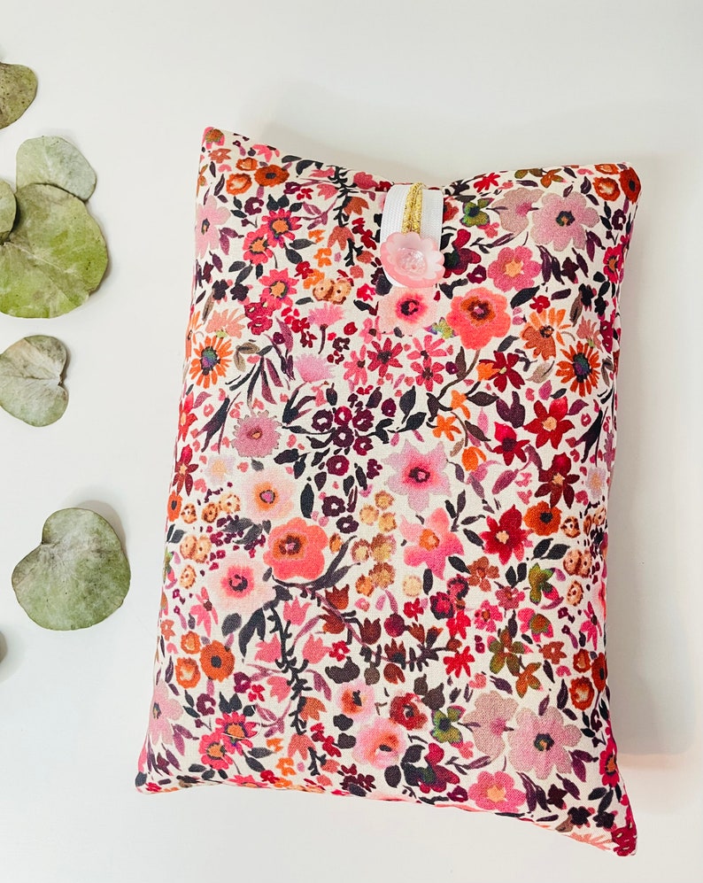 May include: A small, rectangular pillow with a white background and a floral pattern in shades of pink, red, and brown. The pillow has a white ribbon and a pink button.