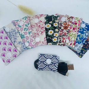 Op de afbeelding: Een verzameling stoffen kabelorganizers in verschillende bloemen- en patroonontwerpen. Elke organizer heeft een drukknoopsluiting. Een blauw-witte organizer met golfpatroon bevat een zwarte kabel en USB-connector.