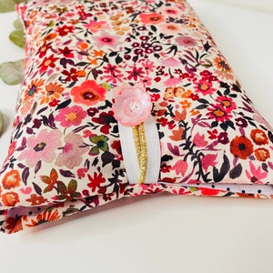 May include: A small, white fabric pouch with a floral print in shades of pink, red, and orange. The pouch has a pink button and a white ribbon with a gold trim.