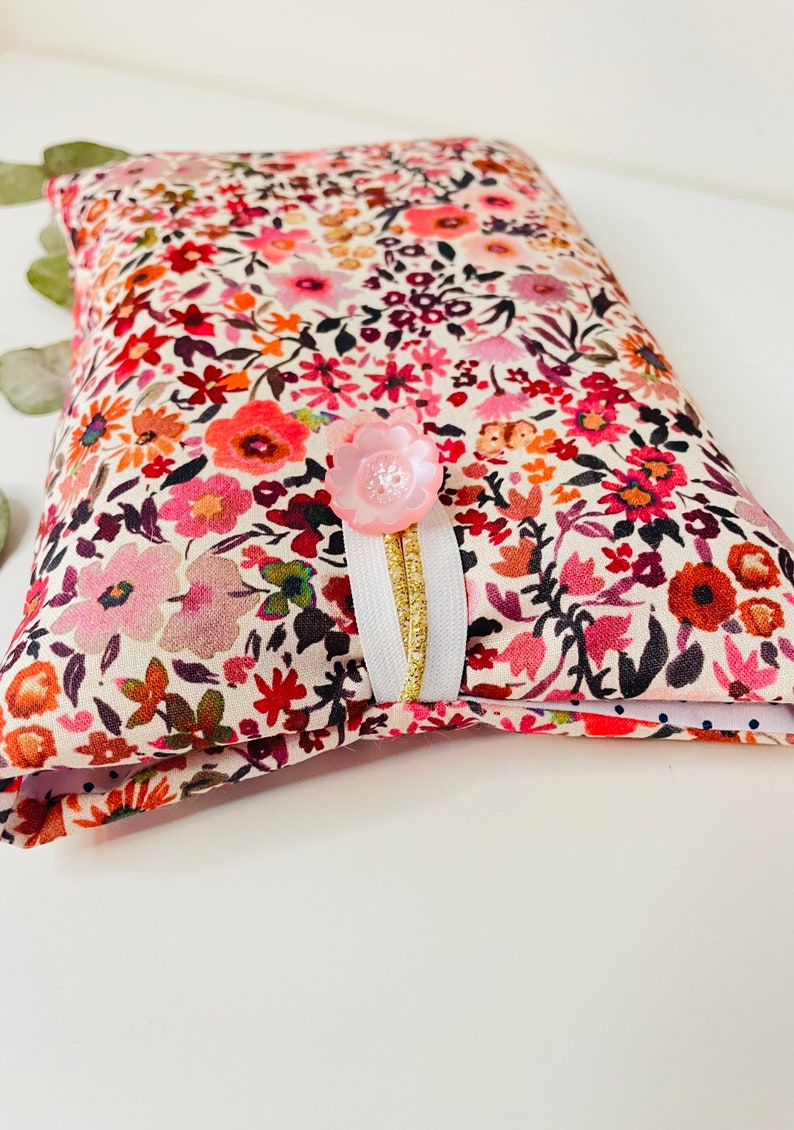 May include: A small, white fabric pouch with a floral print in shades of pink, red, and orange. The pouch has a pink button and a white ribbon with a gold trim.