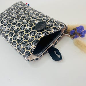 Peut inclure: Une housse de téléphone à motifs géométriques noirs et dorés avec une fermeture à bouton noir. L'étui est ouvert, révélant un téléphone à l'intérieur. Un petit bouquet de fleurs séchées est à côté de l'étui.