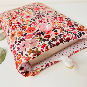 May include: A book cover with a floral pattern in shades of pink, red, and orange. The cover is lined with white fabric with blue polka dots. A pink button is attached to the front of the cover.