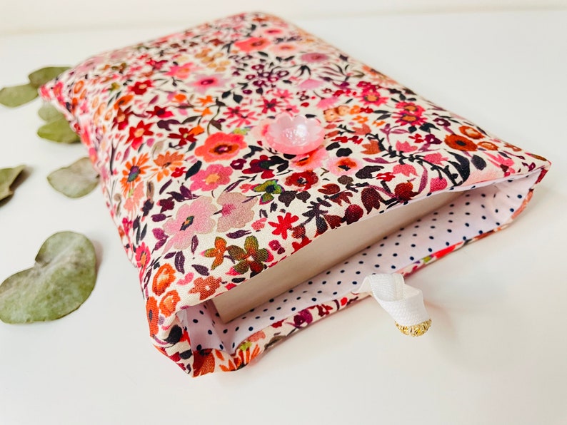 May include: A book cover with a floral pattern in shades of pink, red, and orange. The cover is lined with white fabric with blue polka dots. A pink button is attached to the front of the cover.