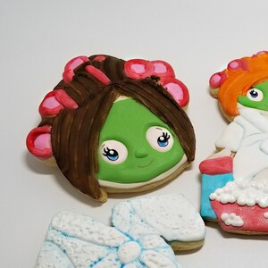 May include: Five decorated sugar cookies with a spa theme. The cookies feature a woman with a green face mask and hair rollers, a woman in a white bathrobe sitting in a tub of bubbles, a tub of bubbles, a towel, and a woman with a green face mask and orange hair.