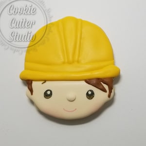 May include: A cookie in the shape of a construction worker's face. The cookie has a yellow hard hat, brown hair, and a smiling face with rosy cheeks. The cookie is likely a themed treat for a construction or building-related event.