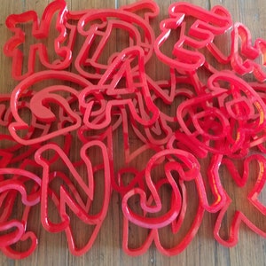 May include: A set of red plastic cookie cutters shaped like letters of the alphabet. The cutters are arranged in a pile, with some letters overlapping.