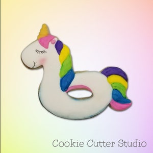 May include: A white unicorn-shaped cookie decorated with rainbow icing, shaped like a pool float. The cookie is a fun and colorful treat for a summer party.