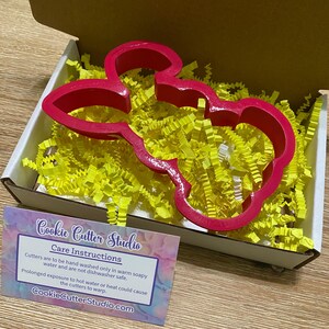May include: A pink plastic cookie cutter shaped like a cartoon bunny rabbit in a box with yellow packing paper. The cutter is for making cookies or fondant decorations. The box has a card with care instructions for the cutter.  Cookie Cutter Studio.com