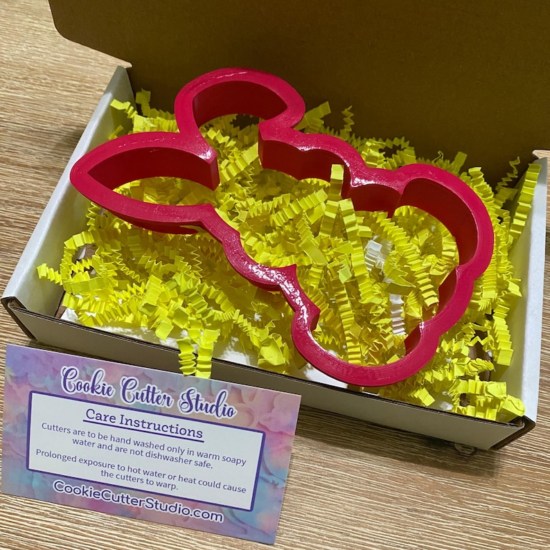 May include: A pink plastic cookie cutter shaped like a cartoon bunny rabbit in a box with yellow packing paper. The cutter is for making cookies or fondant decorations. The box has a card with care instructions for the cutter.  Cookie Cutter Studio.com