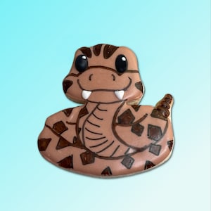 以下が含まれることがあります： ガラガラヘビのデザインで飾られた茶色と黒のクッキー。クッキーは茶色の体に黒い模様があり、白い口に牙があります。クッキーは黒い目と茶色の尻尾をしています。
