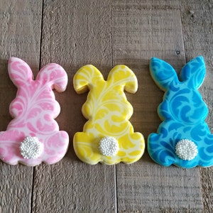 May include: Three decorated sugar cookies shaped like bunnies. The cookies are pink, yellow, and blue with a white swirl design. Each cookie has a white pearl on the bottom.