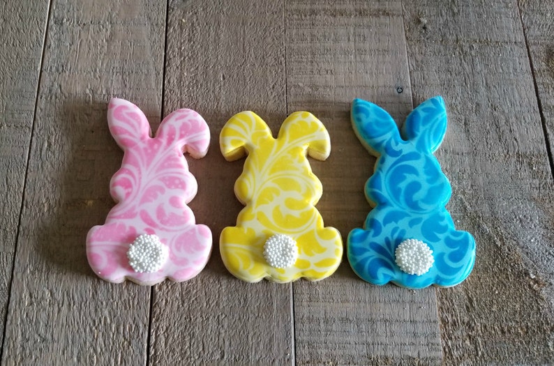 May include: Three decorated sugar cookies shaped like bunnies. The cookies are pink, yellow, and blue with a white swirl design. Each cookie has a white pearl on the bottom.