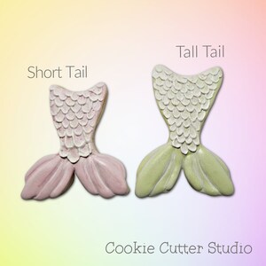 May include: Two mermaid tail cookie cutters, one with a short tail and one with a tall tail. Both cutters are made of white plastic and have a scale pattern. The short tail cutter is pink and the tall tail cutter is green.