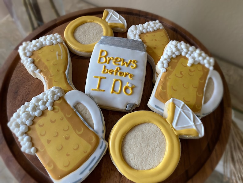 May include: A set of six decorated sugar cookies featuring a beer mug, a beer glass, a ring, and a cookie with the text "Brews before I do".