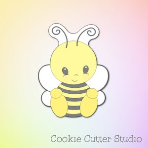 May include: A yellow and black bee cookie cutter with white wings. The bee has a cute cartoon face with gray eyes and a smile. The cookie cutter is designed for making bee-shaped cookies.
