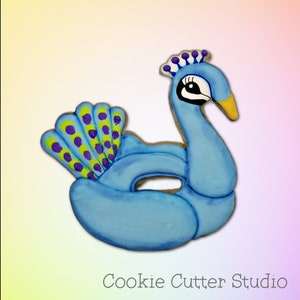May include: A blue peacock-shaped inflatable pool float cookie cutter. The cookie cutter has a detailed peacock design with a purple and yellow tail.