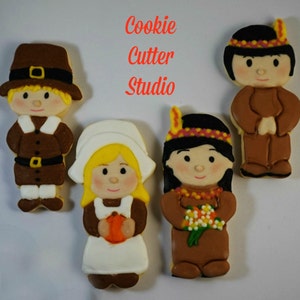 May include: Four decorated sugar cookies depicting a pilgrim girl, a pilgrim boy, a Native American girl, and a Native American boy. The cookies are decorated with icing and feature details like hats, feathers, and flowers. The cookies are arranged on a white surface. Cookie Cutter Studio is written in red text at the top of the image.