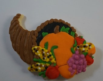 May include: A brown and white cornucopia cookie decorated with a pumpkin, grapes, corn, and red tomatoes.
