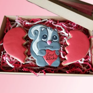 May include: A gray and white skunk cookie holding a red heart cookie that says "Love Stinks". The skunk cookie is in a box with red and white shredded paper. There are two broken red heart cookies in the box.