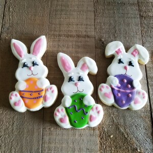 May include: Three decorated sugar cookies shaped like bunnies holding Easter eggs. The bunny on the left is holding an orange egg with purple stripes. The bunny in the middle is holding a green egg with a white swirl. The bunny on the right is holding a purple egg with white polka dots.