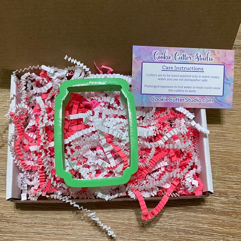 May include: Green plastic cookie cutter shaped like a can of soda. The cutter is in a box with pink and white shredded paper. The box also includes a card with the text "Cookie Cutter Studio" and care instructions for the cutter.