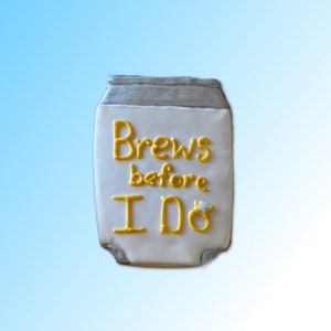 May include: A white iced cookie shaped like a can of beer with the words "Brews before I Do" in yellow icing.