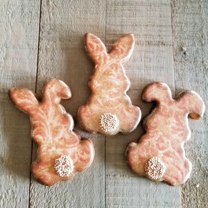 May include: Three pink and gold bunny-shaped sugar cookies decorated with white sprinkles. The cookies are arranged on a wooden surface.