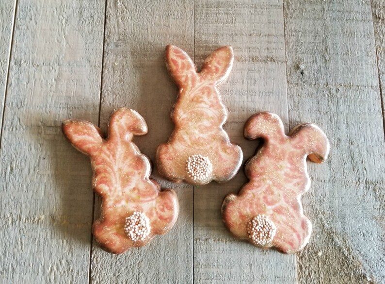 May include: Three pink and gold bunny-shaped sugar cookies decorated with white sprinkles. The cookies are arranged on a wooden surface.