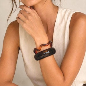 May include: Two brown and black bangles are worn on the arm of a person wearing a white sleeveless top. The top is paired with white pants and a brown belt. Gold hoop earrings are also visible.