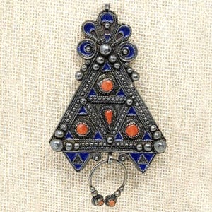 May include: A triangular silver pendant featuring intricate metalwork, deep blue enamel detailing, and four small round orange stones. The piece is topped with a decorative floral-shaped element and includes a small circular ring at the bottom.