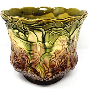 May include: A glazed ceramic planter featuring a scalloped rim and a relief pattern of swirling leaves and vines. The surface displays a gradient of colors, transitioning from a deep brown at the base to a vibrant olive green at the top edge.