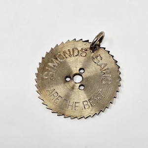 May include: Circular metal pendant resembling a saw blade. The pendant has a ring for attachment and features the engraved text "SIMONDS SAWS ARE THE BEST". The blade has a series of teeth around the edge.