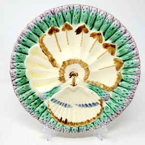 May include: A round ceramic serving dish featuring a cream-colored center with brown painted floral details and a scalloped rim decorated with textured green and purple leaves. The dish has a divided interior design with a central handle element.