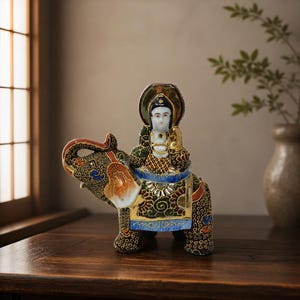 May include: A ceramic figurine featuring a figure with a white face and gold robes seated atop a dark elephant. The elephant is decorated with intricate gold, green, blue, and red patterns, including floral motifs and a saddle blanket with gold trim.